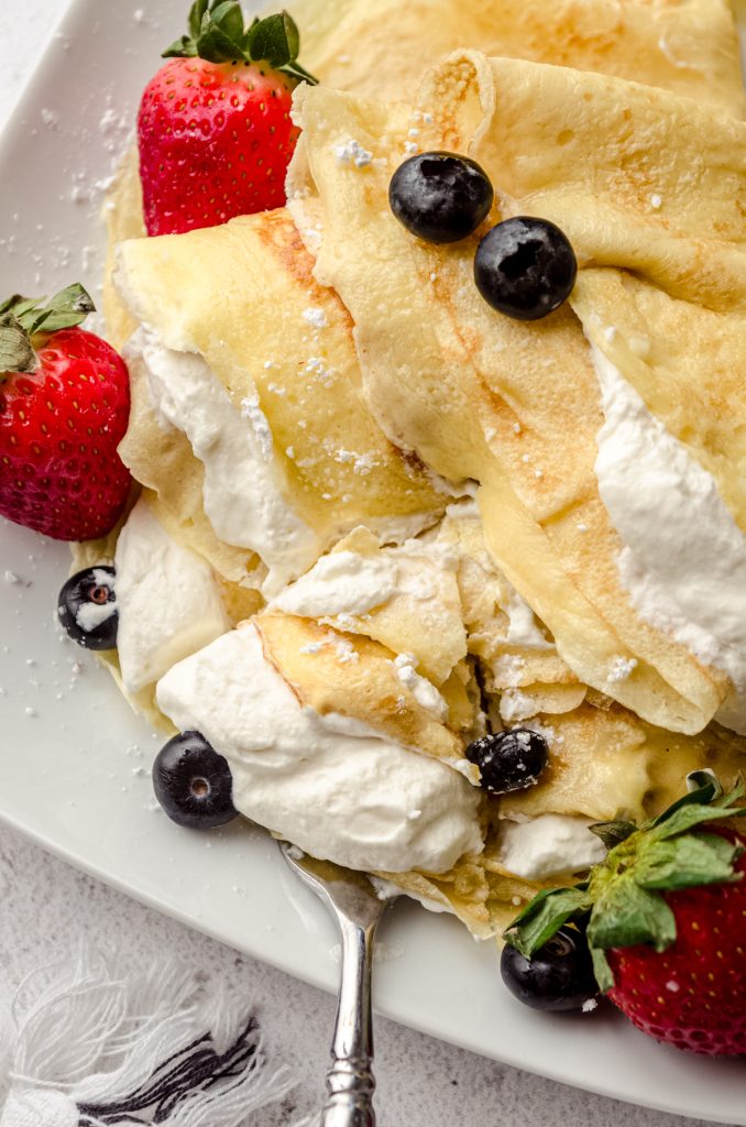 A photo of crepes on a plate with fresh whipped cream, blueberries, and strawberries on them and a fork taking a triangle shaped piece out of the stack.