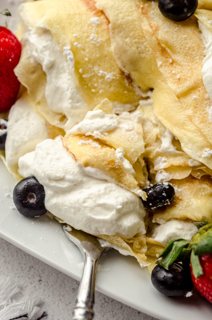 A photo of crepes on a plate with fresh whipped cream, blueberries, and strawberries on them and a fork taking a triangle shaped piece out of the stack.