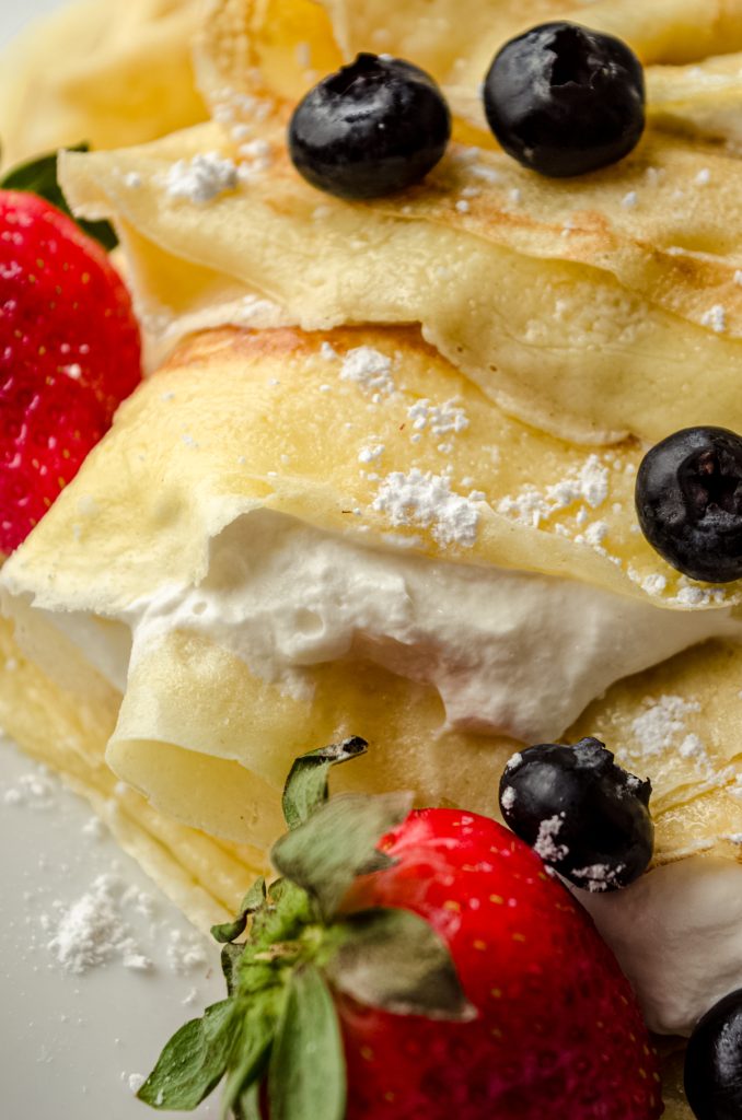 A photo of crepes on a plate with fresh whipped cream, blueberries, and strawberries on them.