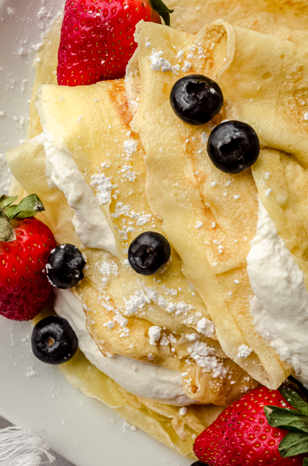 A photo of crepes on a plate with fresh whipped cream, blueberries, and strawberries on them.