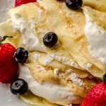 A photo of crepes on a plate with fresh whipped cream, blueberries, and strawberries on them.