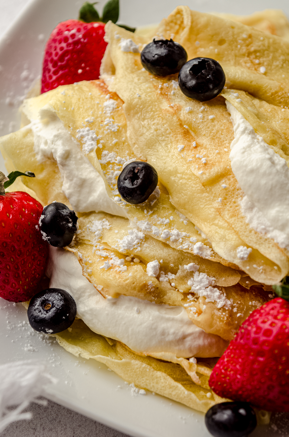 A photo of crepes on a plate with fresh whipped cream, blueberries, and strawberries on them.