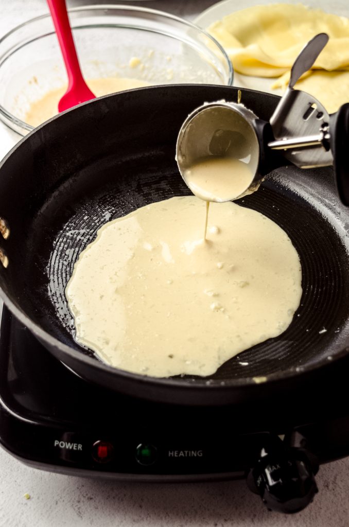 Someone is using an ice cream scoop to pour crepe batter into a skillet.