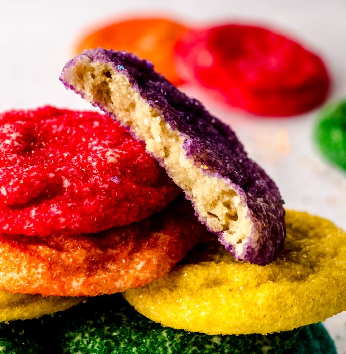 A stack of rainbow colored sugar cookies with a purple one on top that has been cut in half to see the cross-section.