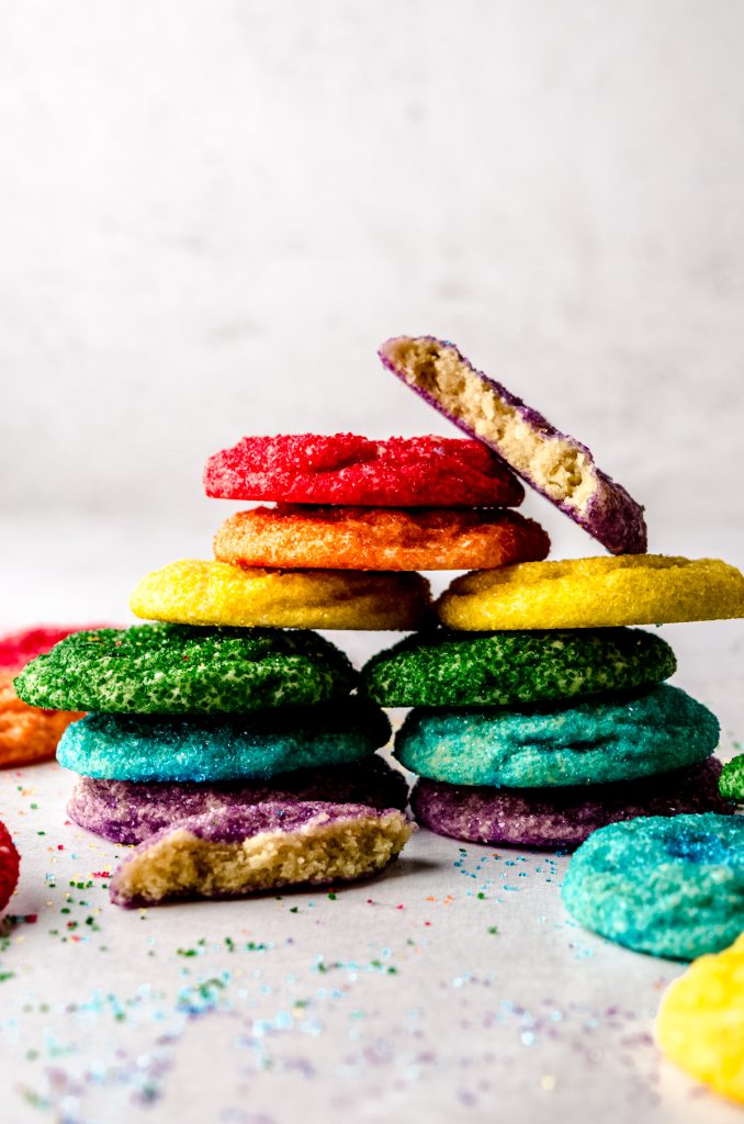 A stack of rainbow colored sugar cookies with a purple one on top that has been cut in half to see the cross-section.