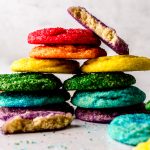 A stack of rainbow colored sugar cookies with a purple one on top that has been cut in half to see the cross-section.