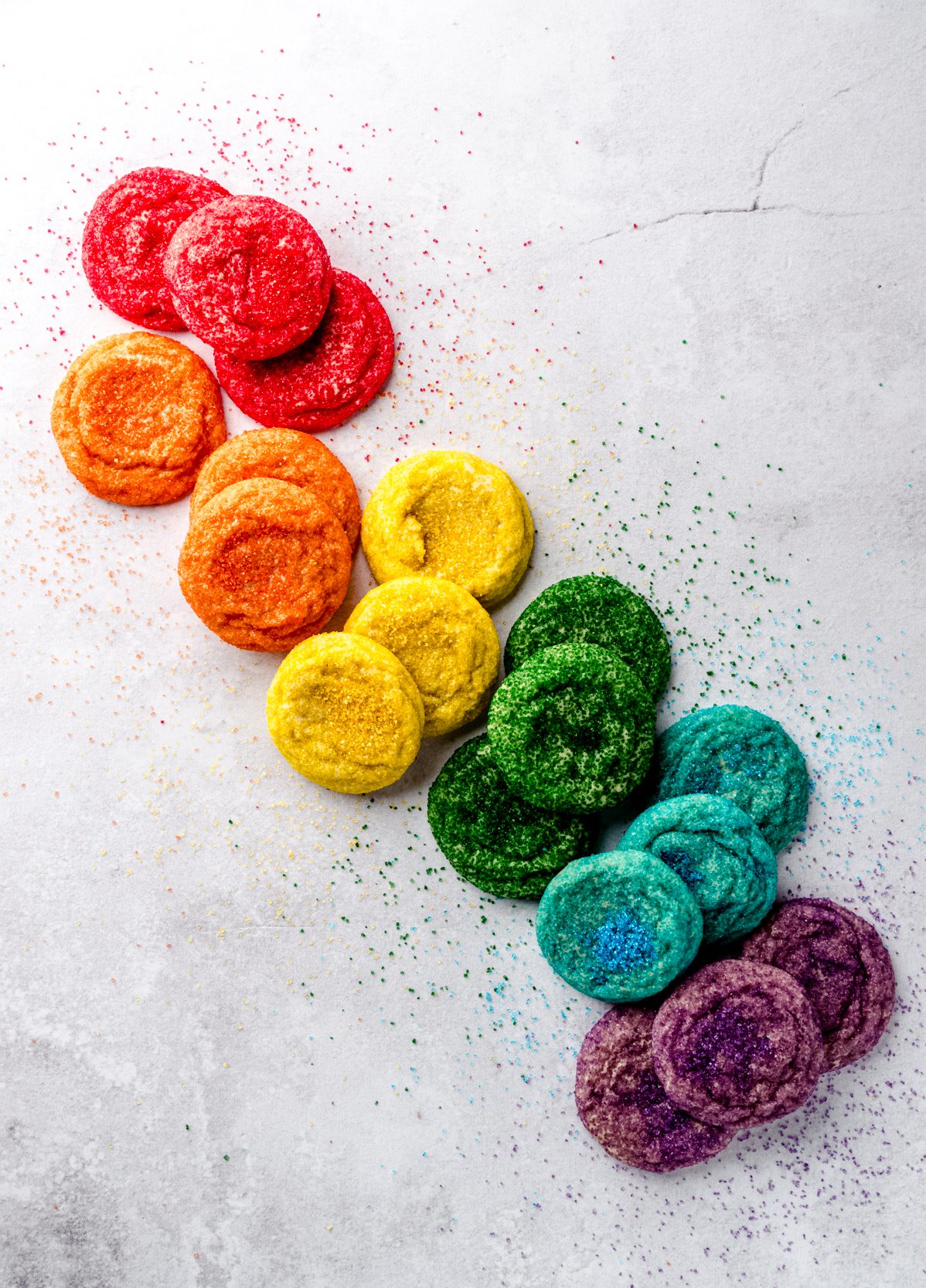 An aerial photo of sugar cookies coated in red, orange, yellow, green, blue, and purple sugar arranged on a surface in color order.