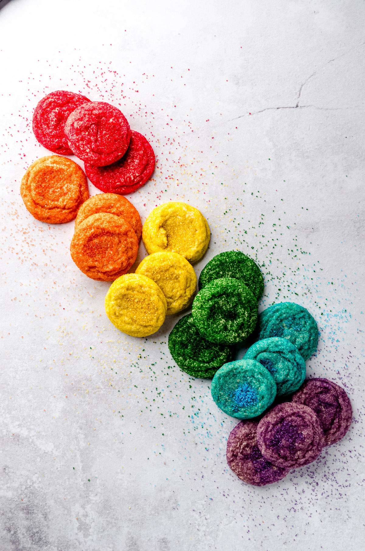 An aerial photo of sugar cookies coated in red, orange, yellow, green, blue, and purple sugar arranged on a surface in color order.