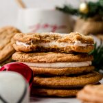 A stack of gingerbread sandwich cookies on a plate with a bite taken out of the one on the top and you can see a mug and a Christmas tree in the background.