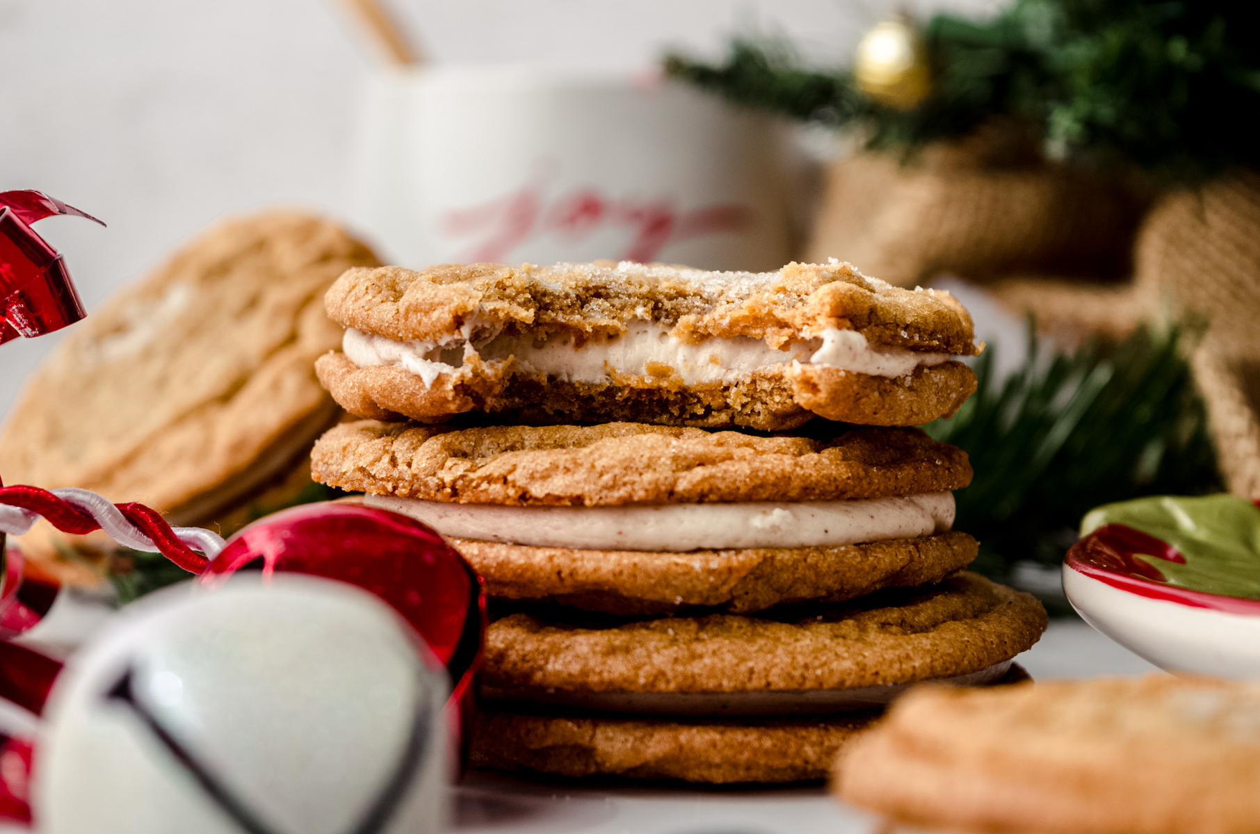 A stack of gingerbread sandwich cookies on a plate with a bite taken out of the one on the top and you can see a mug and a Christmas tree in the background.