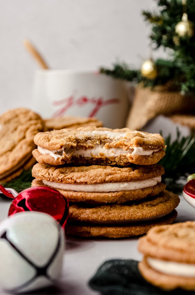 A stack of gingerbread sandwich cookies on a plate with a bite taken out of the one on the top and you can see a mug and a Christmas tree in the background.