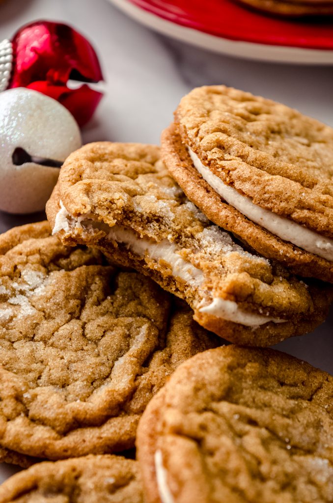 Gingerbread sandwich cookies on a surface and you can see Christmas bells and other decor in the background and a bite has been taken out of one of the sandwich cookies.