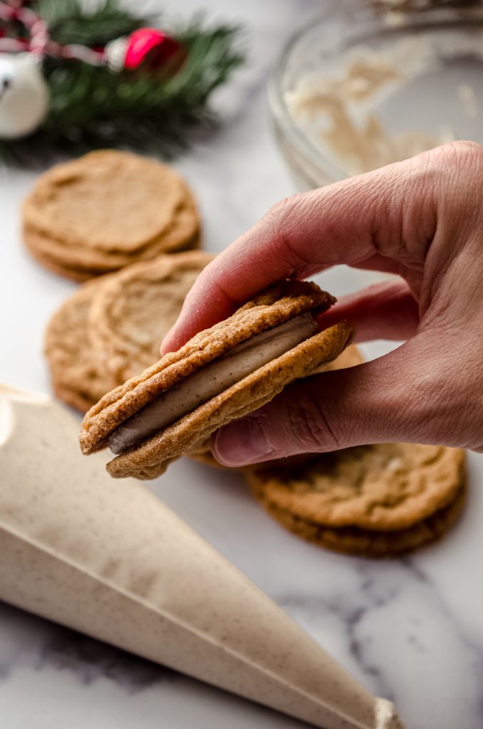 Someone is holding a gingerbread sandwich cookie and you can see more cookies, a piping bag with frosting, and Christmas decor in the background.