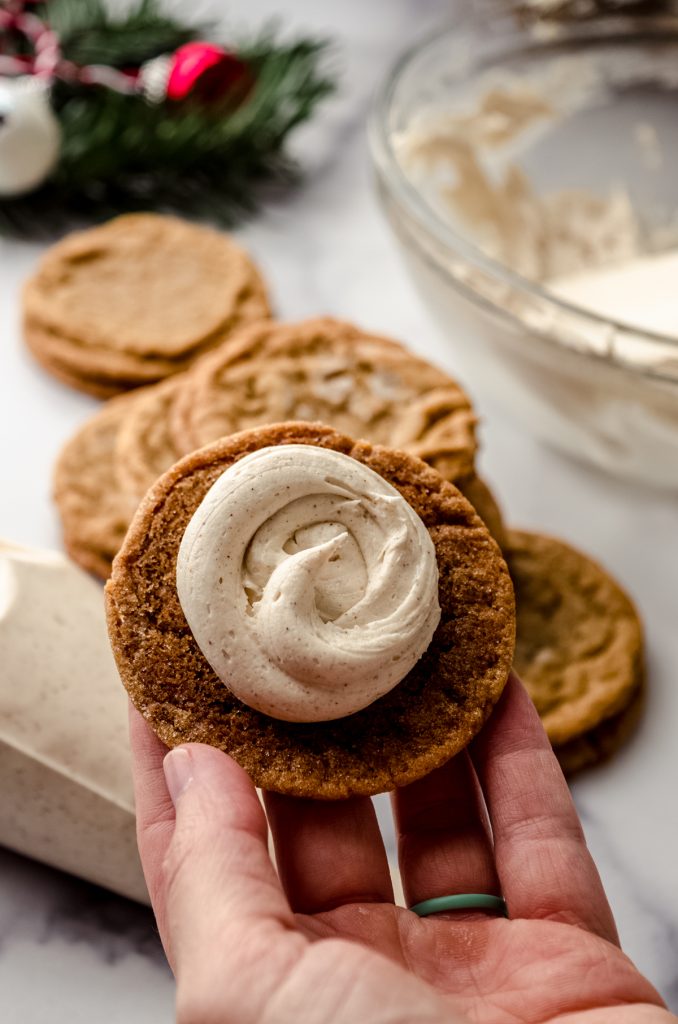 Someone is holding a gingerbread cookie, bottom side up, with frosting piped onto it, and you can see more cookies, a bowl of frosting, and Christmas decor in the background.