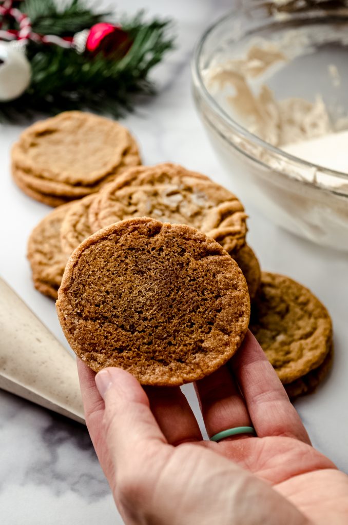 Someone is holding a gingerbread cookie, bottom side up, and you can see more cookies, a bowl of frosting, and Christmas decor in the background.