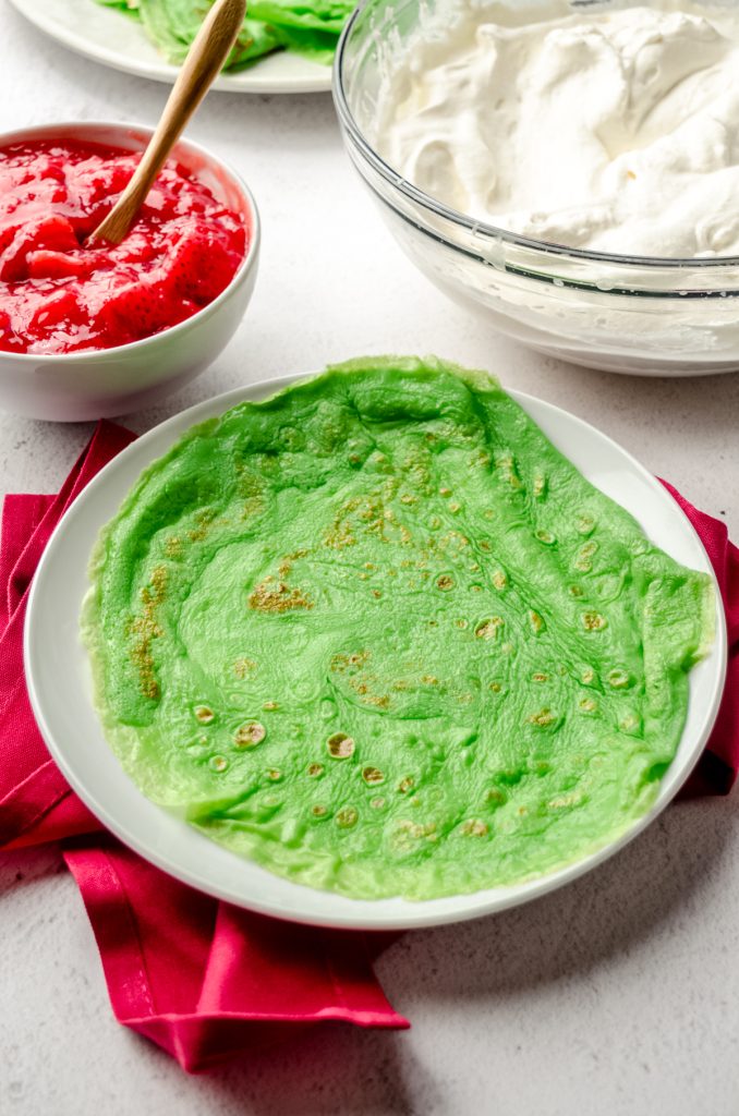 A green crepe on a plate with strawberry sauce and whipped cream in the background.