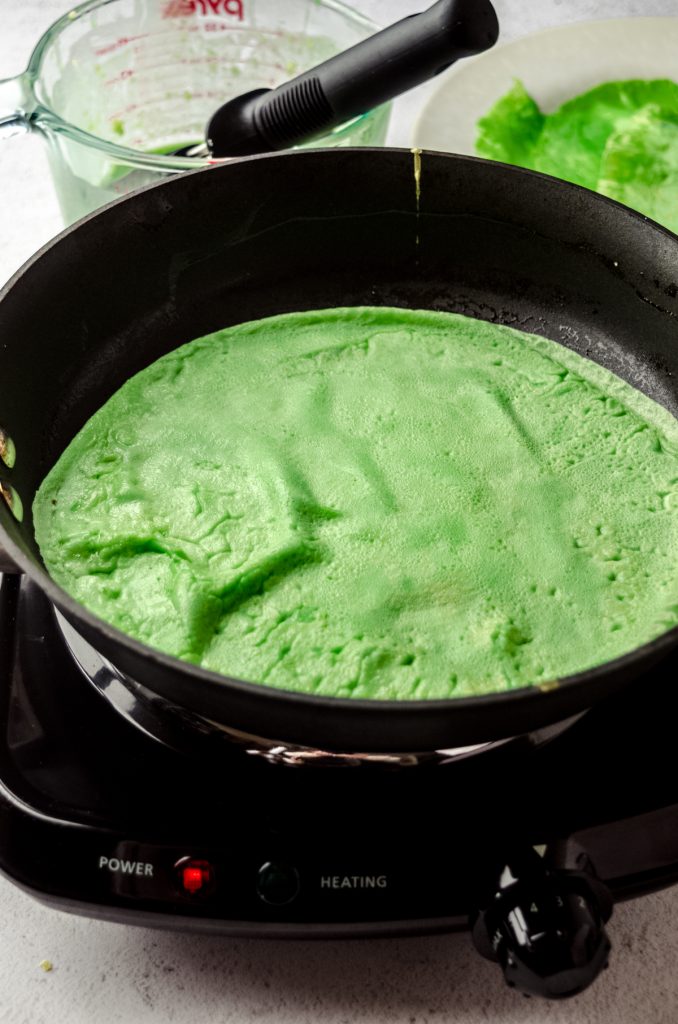 A green crepe cooking in a pan in preparation of making Grinch crepes.