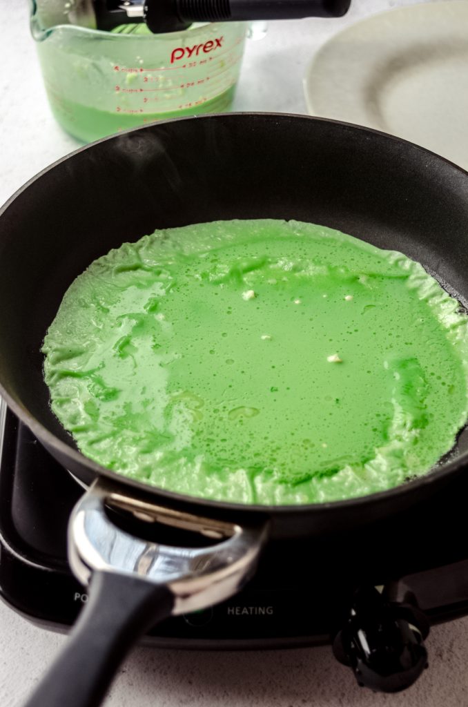 A green crepe cooking in a pan in preparation of making Grinch crepes.