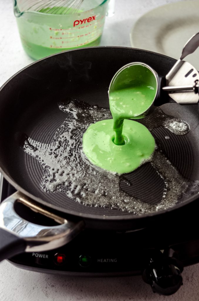 Someone is using a scoop to pour green batter into a buttered pan to make Grinch crepes.