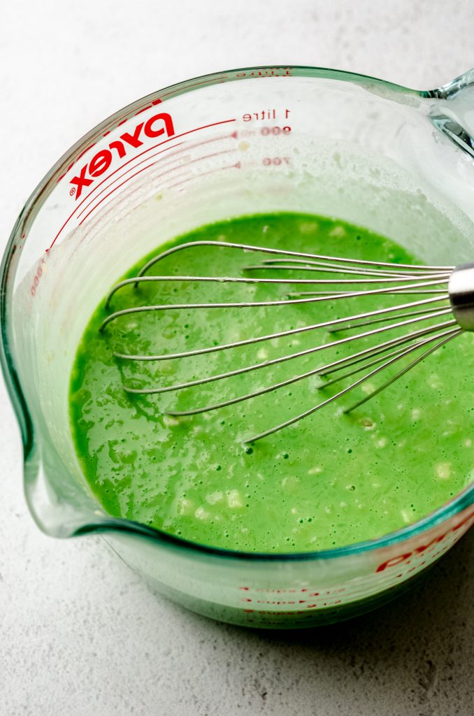 Green crepe batter in a large measuring cup with a whisk.