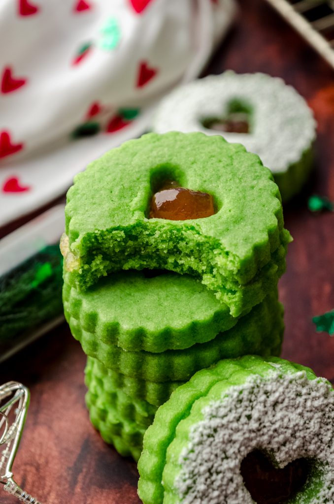 A stack of Grinch Linzer cookies on a surface and a bite has been taken out of the one on the top.