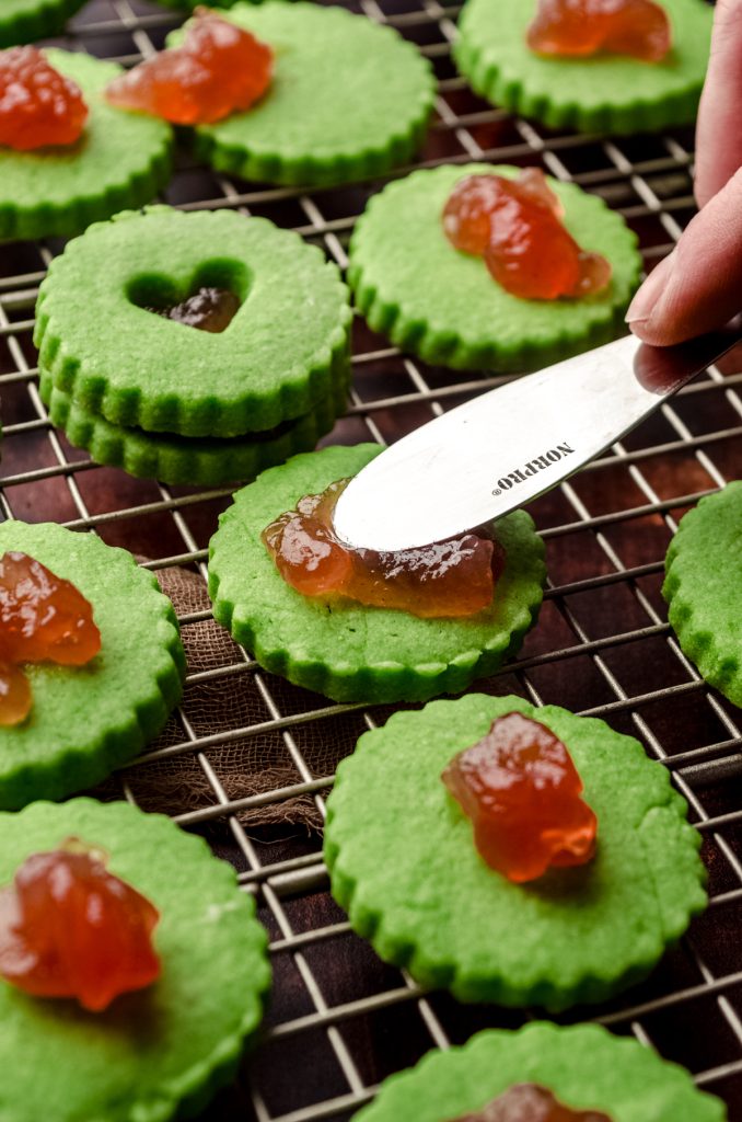 Someone is spreading the filling on the bottom of a Grinch Linzer sandwich cookie.