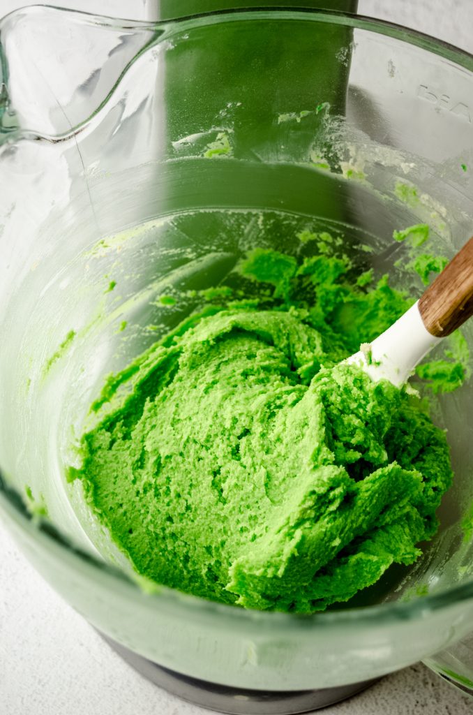 Green Linzer cookie dough in the bowl of a mixer with a spatula in it.