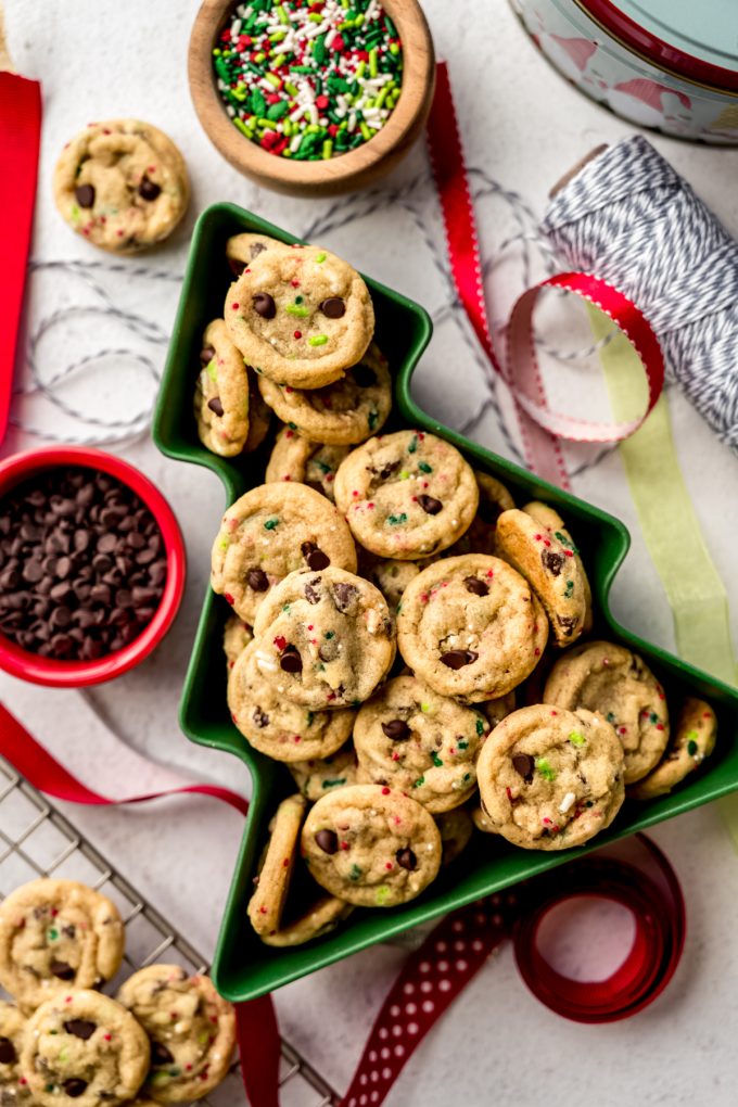 Aerial photo of mini Christmas chocolate chip cookies with festive sprinkles in it sitting in a tree shaped bowl with Christmas ribbon and a small bowl of chocolate chips around it.