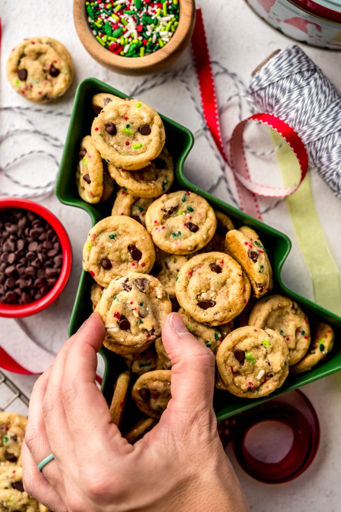 Aerial photo of mini Christmas chocolate chip cookies sitting in a tree shaped dish and someone is grabbing one of the cookies.