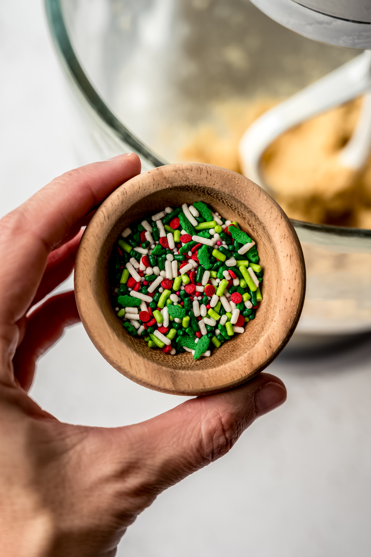 Someone's hand holding a small wooden bowl filled with Christmas sprinkles.