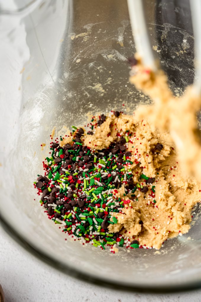 Christmas chocolate chip cookie dough in the bowl of a stand mixer with sprinkles and chocolate chips added to it before getting blended in.