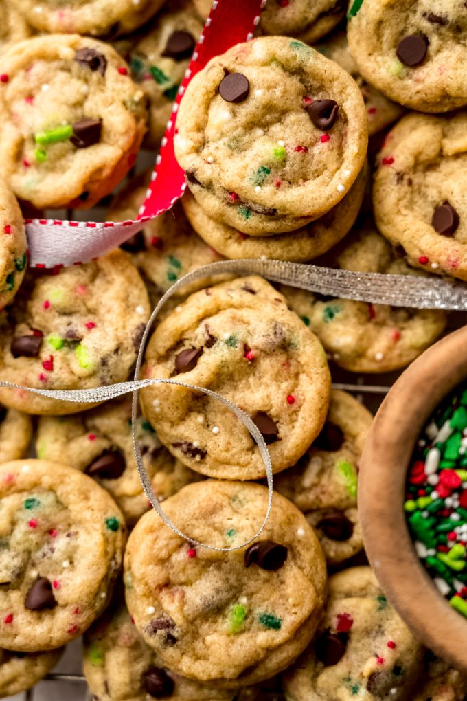 Aerial photo of mini Christmas chocolate chip cookies on a surface with festive ribbon and a bowl of sprinkles around it.