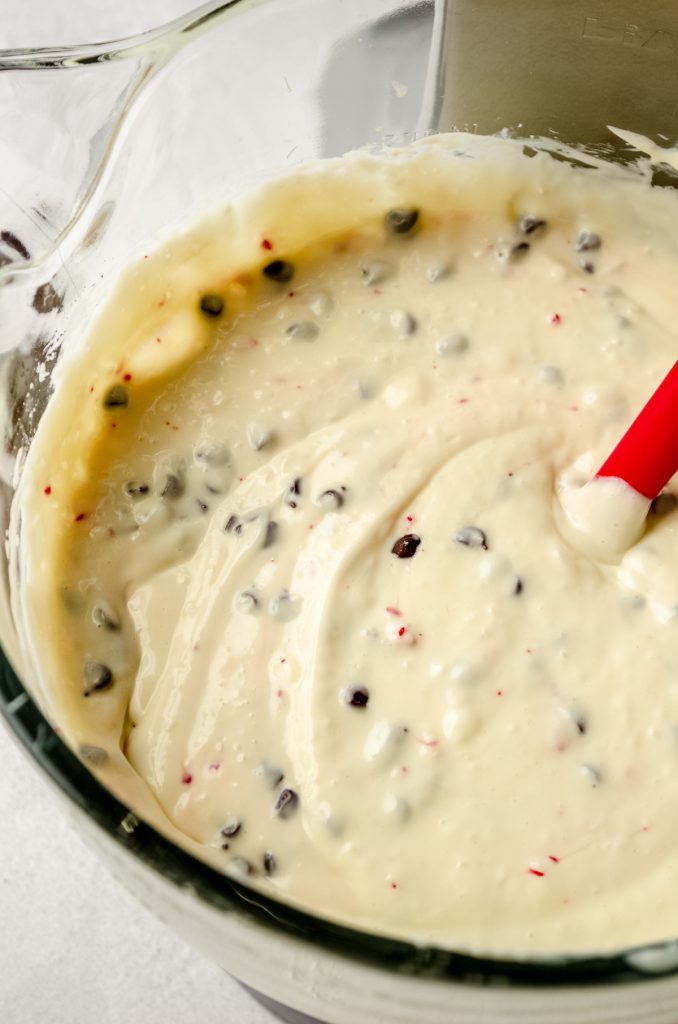 Peppermint bark cheesecake batter in the bowl of a mixer with a red spatula in it.