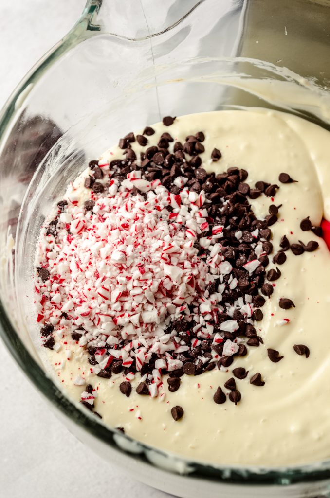 Peppermint bark cheesecake batter in the bowl of a mixer with a red spatula in it getting ready to stir in the chocolate chips and candy cane bits.