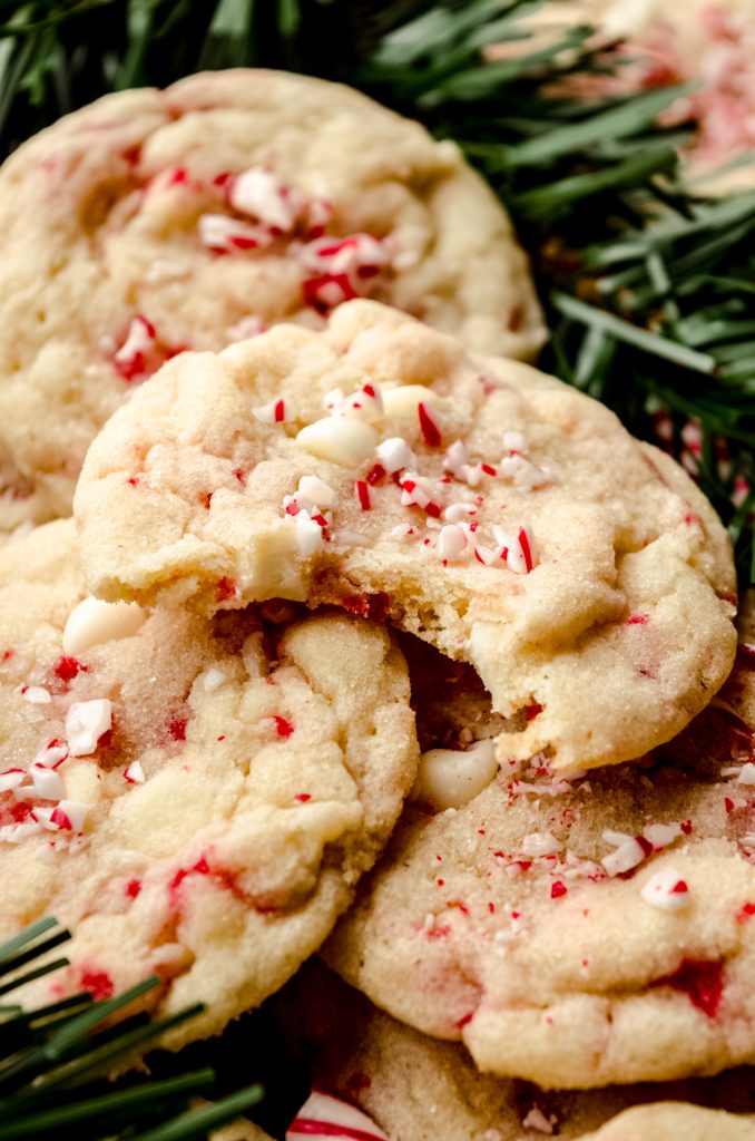 White chocolate chip peppermint cookies on a surface with candy cane pieces and evergreen needles around it and a bite taken out of the cookie on the top.