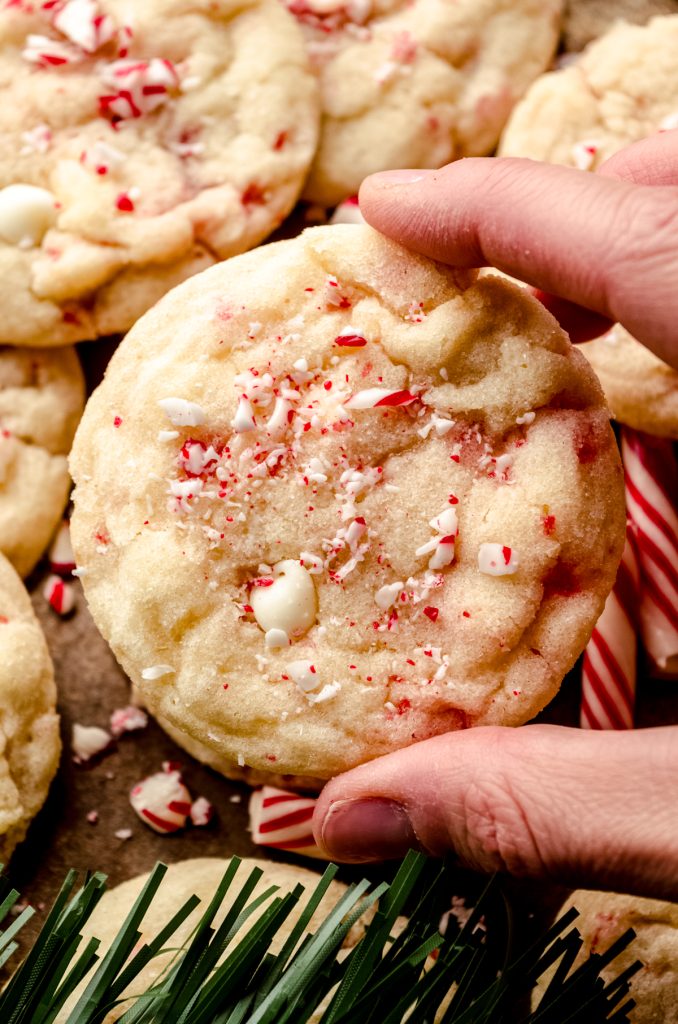 Someone's hand holding a white chocolate chip peppermint cookie on a surface with candy cane pieces around it and evergreen needles in the foreground.