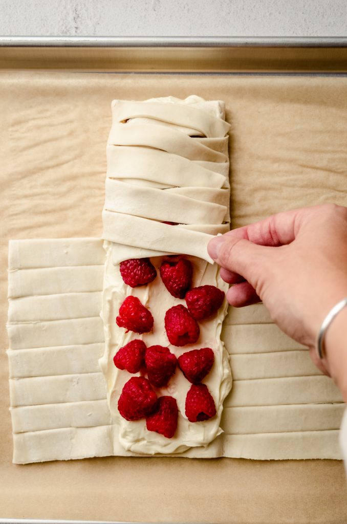 Someone is folding the strips of a puff pastry filled with cream cheese and raspberries to make a pastry braid.