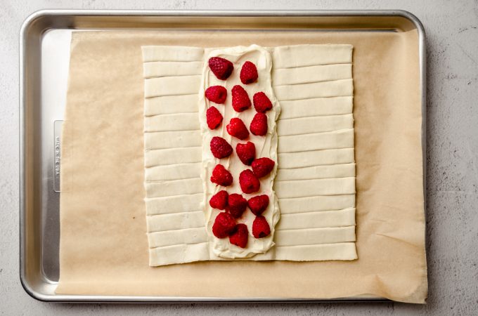 A rectangle of puff pastry dough on a baking sheet lined with parchment paper with cream cheese filling and fresh raspberries on the center flap.