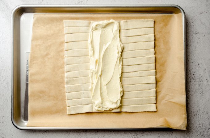 A rectangle of puff pastry dough on a baking sheet lined with parchment paper with cream cheese filling on the center flap.