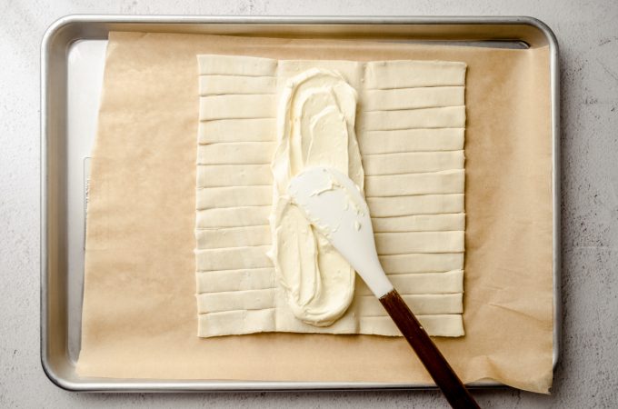 A rectangle of puff pastry dough on a baking sheet lined with parchment paper and someone is spreading a cream cheese filling into the center flap.