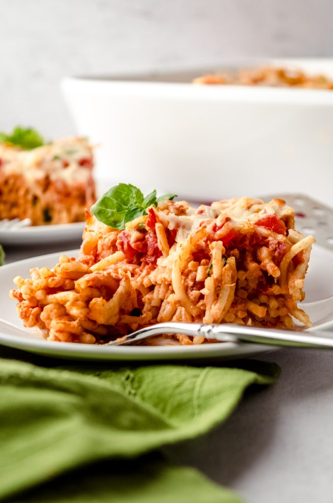 A slice of baked cream cheese spaghetti casserole on a plate with a fork that has a bite of the casserole on it.