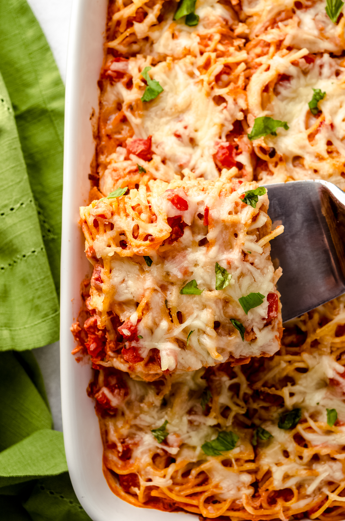 Someone is lifting a slice of baked cream cheese spaghetti casserole out of a baking dish.