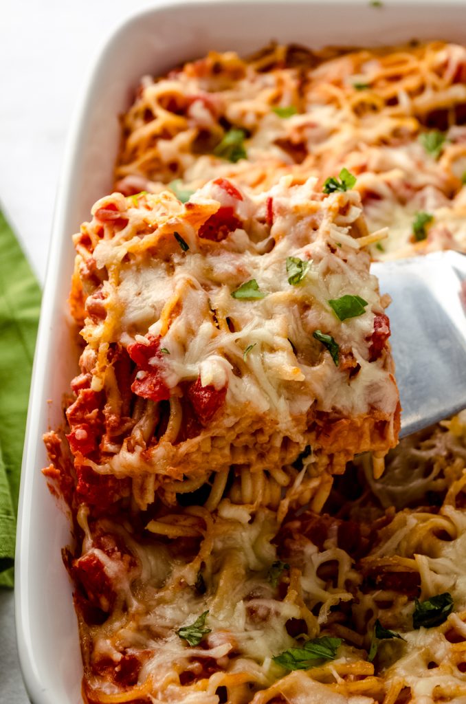 Someone is lifting a slice of baked cream cheese spaghetti casserole out of a baking dish.