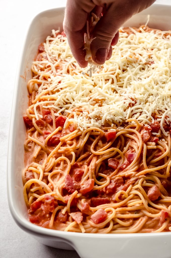 Someone's hand sprinkling shredded mozzarella onto a spaghetti casserole.