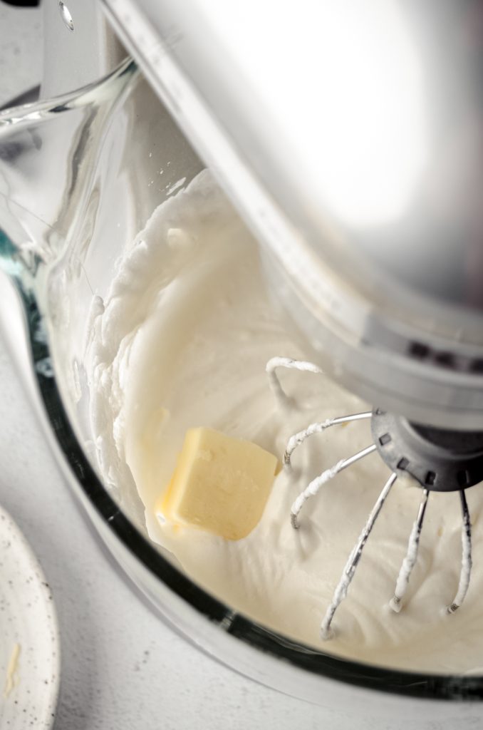 The bowl of a stand mixer fitted with the whisk attachment with meringue in the bowl and a pat of butter.