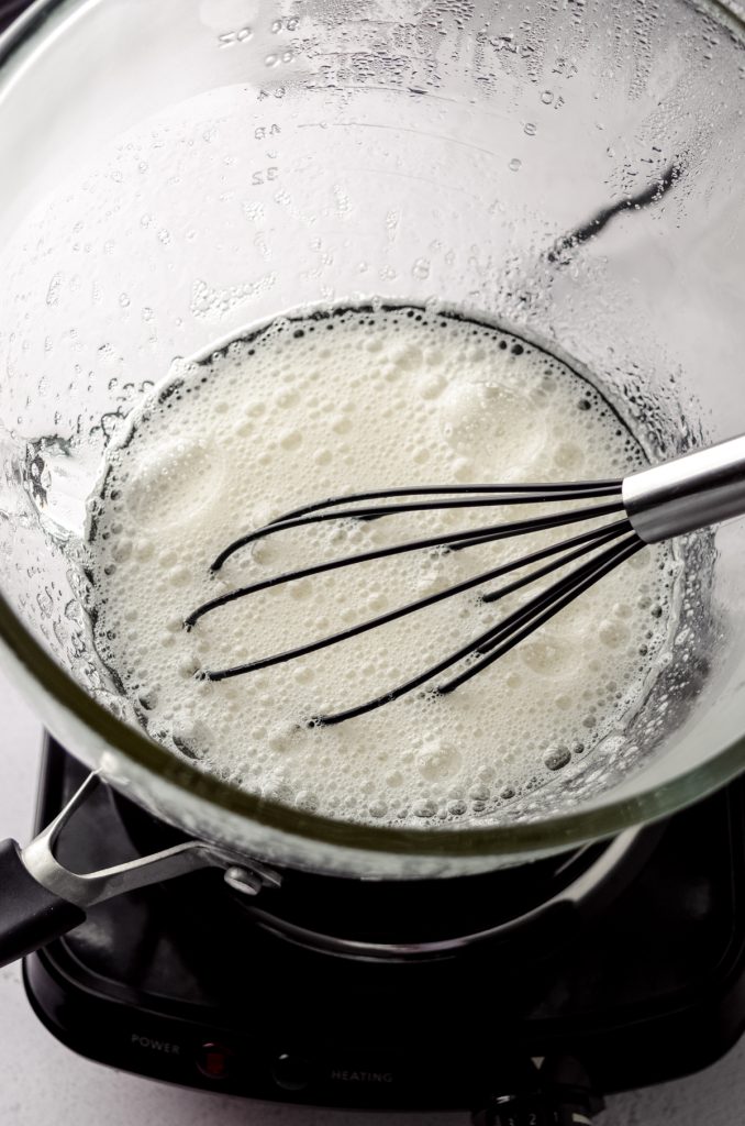 A glass bowl over a pot of simmering water to create a double boiler with egg whites, sugar, and a whisk in it.