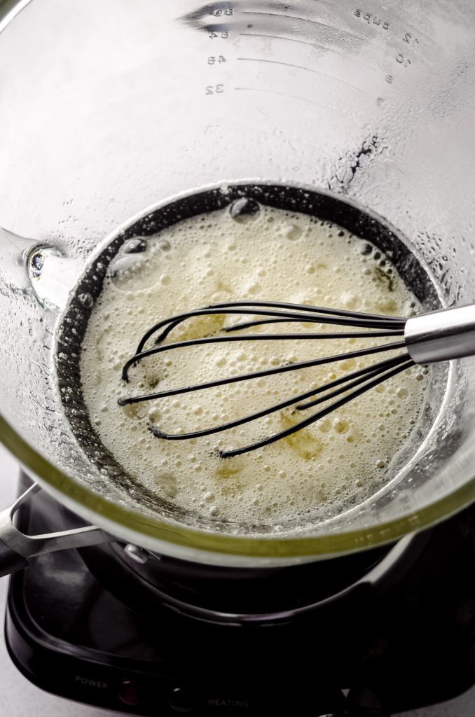 A glass bowl over a pot of simmering water to create a double boiler with egg whites, sugar, and a whisk in it.