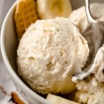 A bowl of roasted banana ice cream in a bowl with a spoon, banana slices, and pieces of waffle cone in it.