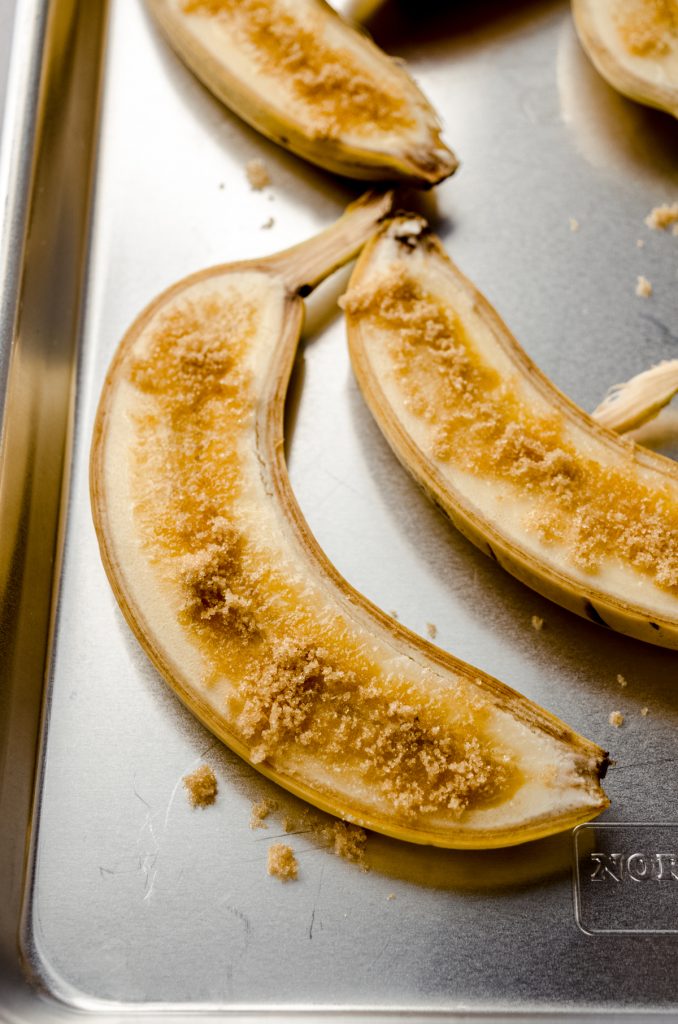 Yellow bananas that have been sliced long ways and sprinkled with brown sugar lying face up on a baking sheet.