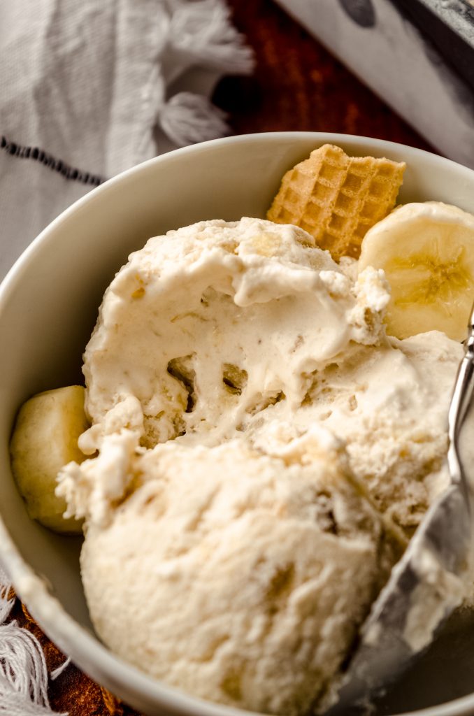 A bowl of roasted banana ice cream in a bowl with a spoon, banana slices, and pieces of waffle cone in it.